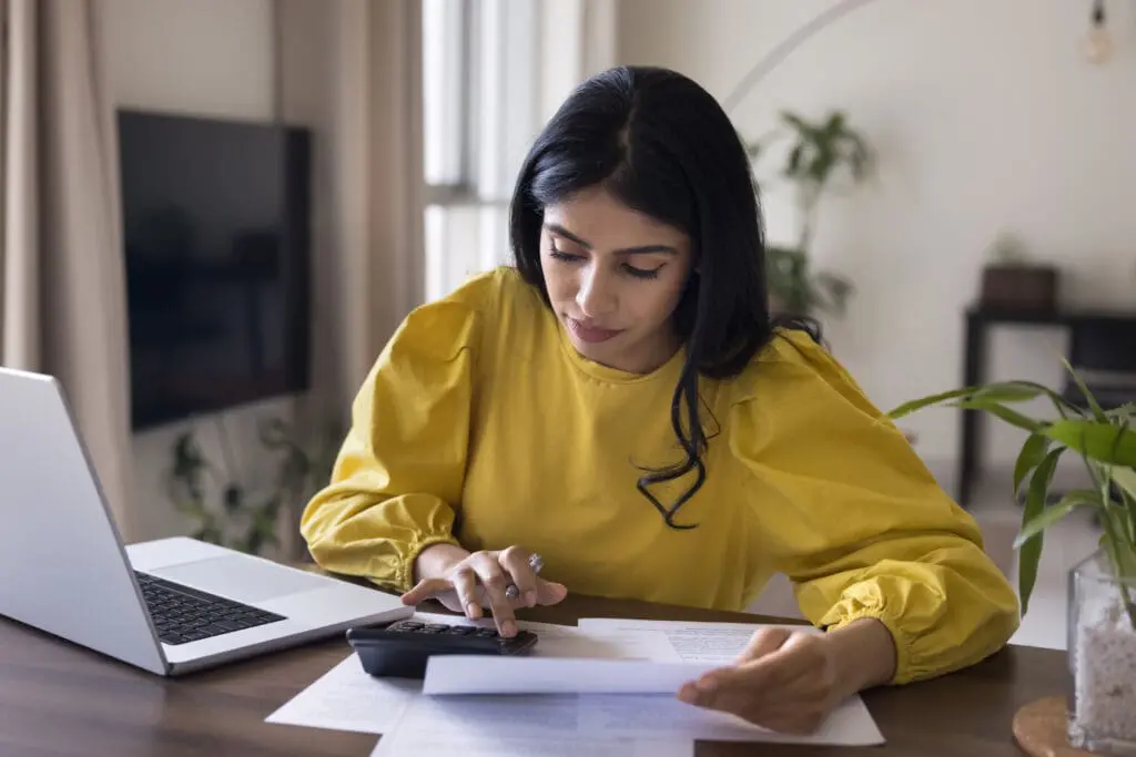 Focused busy Indian freelance accountant woman checking financial documents, counting budget, income, expenses on calculator, sitting at home workplace table with laptop.